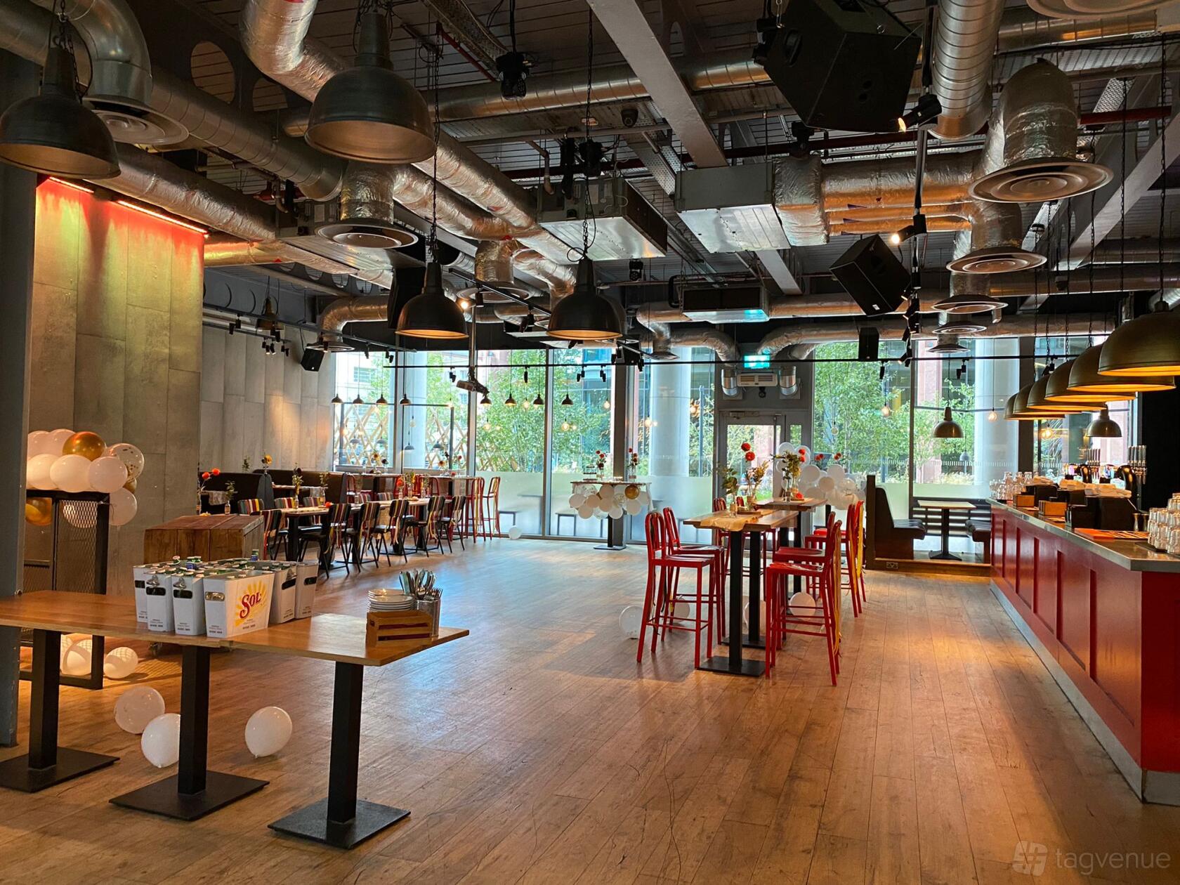 A bar with industrial-style exposed ductwork, wood floors, large windows, and high tables at The Union Regents.