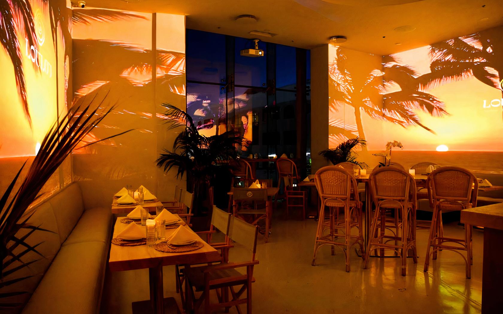 A restaurant dining room with warm sunset lighting, palm tree projections, and rattan chairs at LouLou.