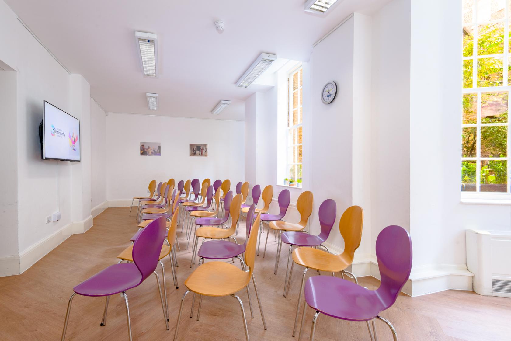 A function room with rows of purple and yellow chairs, wood flooring, large windows, and a wall-mounted screen at Fitzrovia Community Centre.