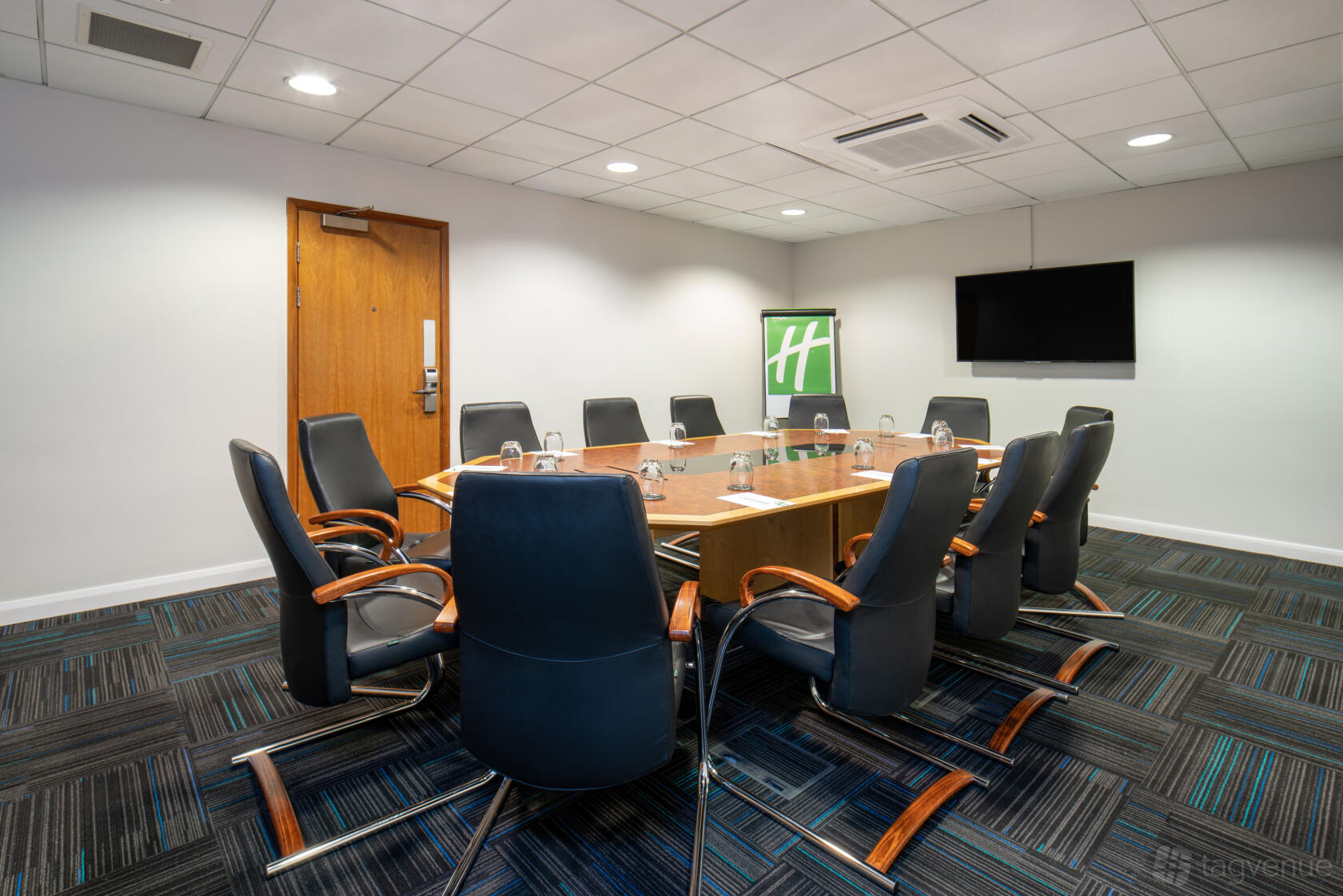 A boardroom with a wooden conference table, leather chairs, wall-mounted screen, and carpet flooring at Holiday Inn London - Brentford Lock.