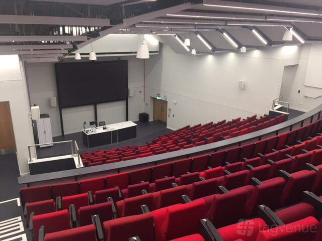 An auditorium with tiered rows of red seats, large projection screen, and lecture podium at ARU Venue Hire - Cambridge.