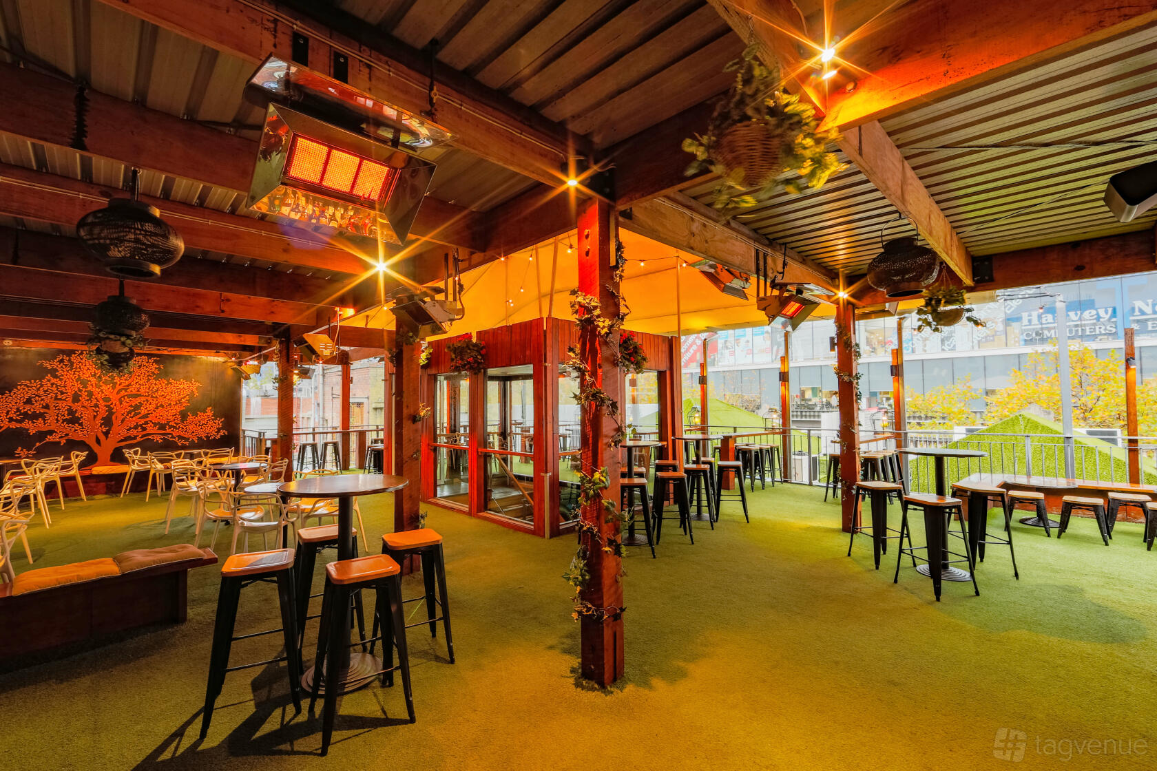 A rooftop bar with artificial grass flooring, wooden beams, hanging plants, and high tables at The Emerald Peacock.