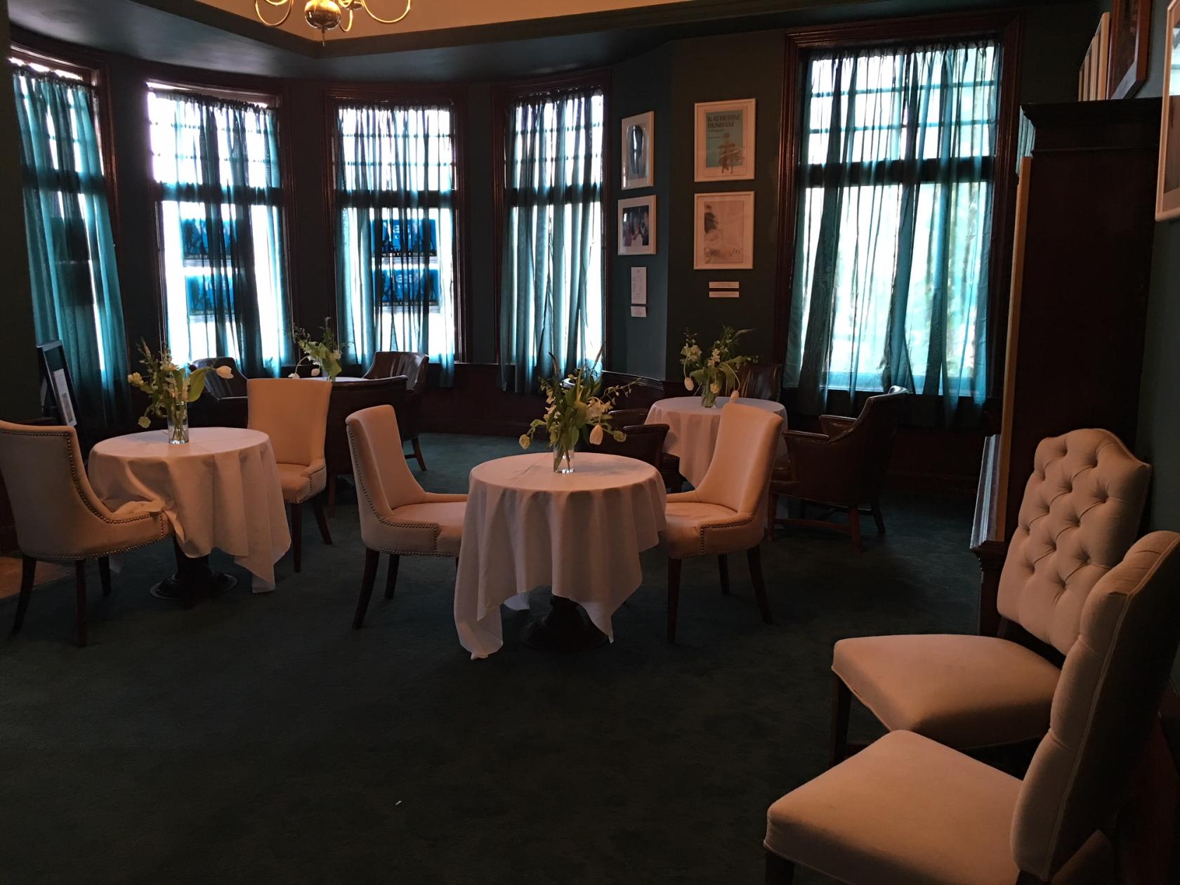 A hall with round tables draped in white linens and cream upholstered chairs by large windows at Geoffrey's Inner Circle.
