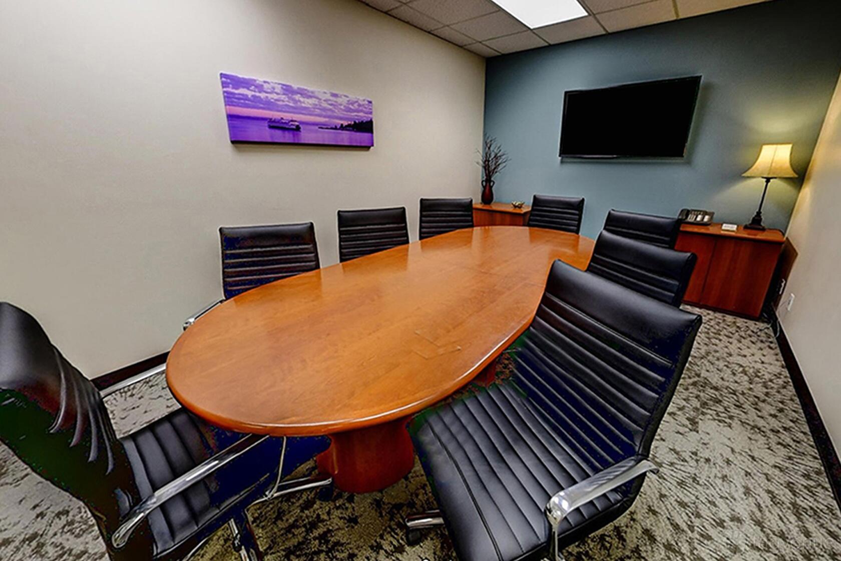 A meeting room with an oval wooden conference table, black leather chairs, wall-mounted TV, and a table lamp at Premier Workspaces - Northcreek.