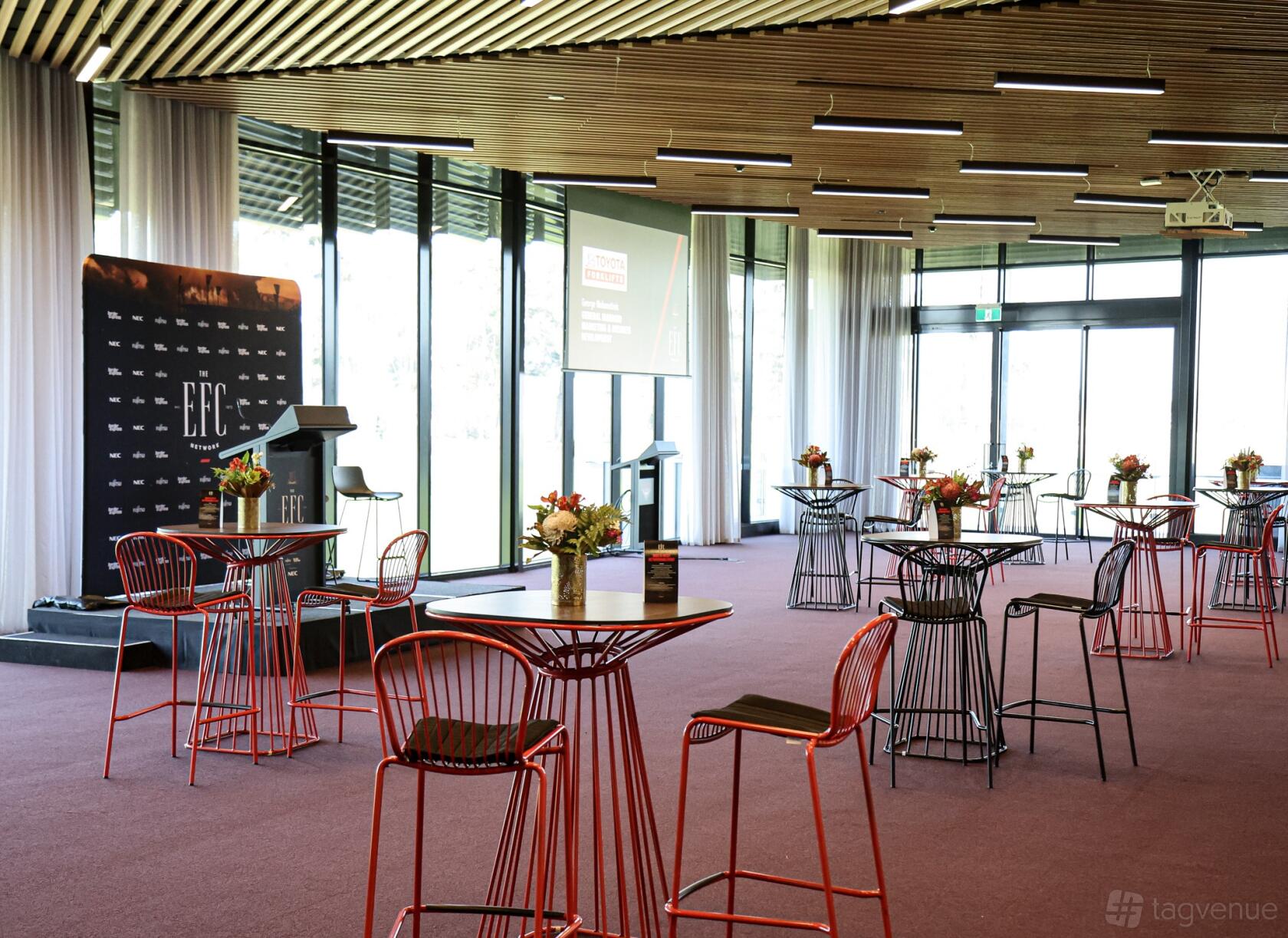 A function room with floor-to-ceiling windows, round cocktail tables with red chairs, and a podium at NEC Hangar - Essendon Football Club.