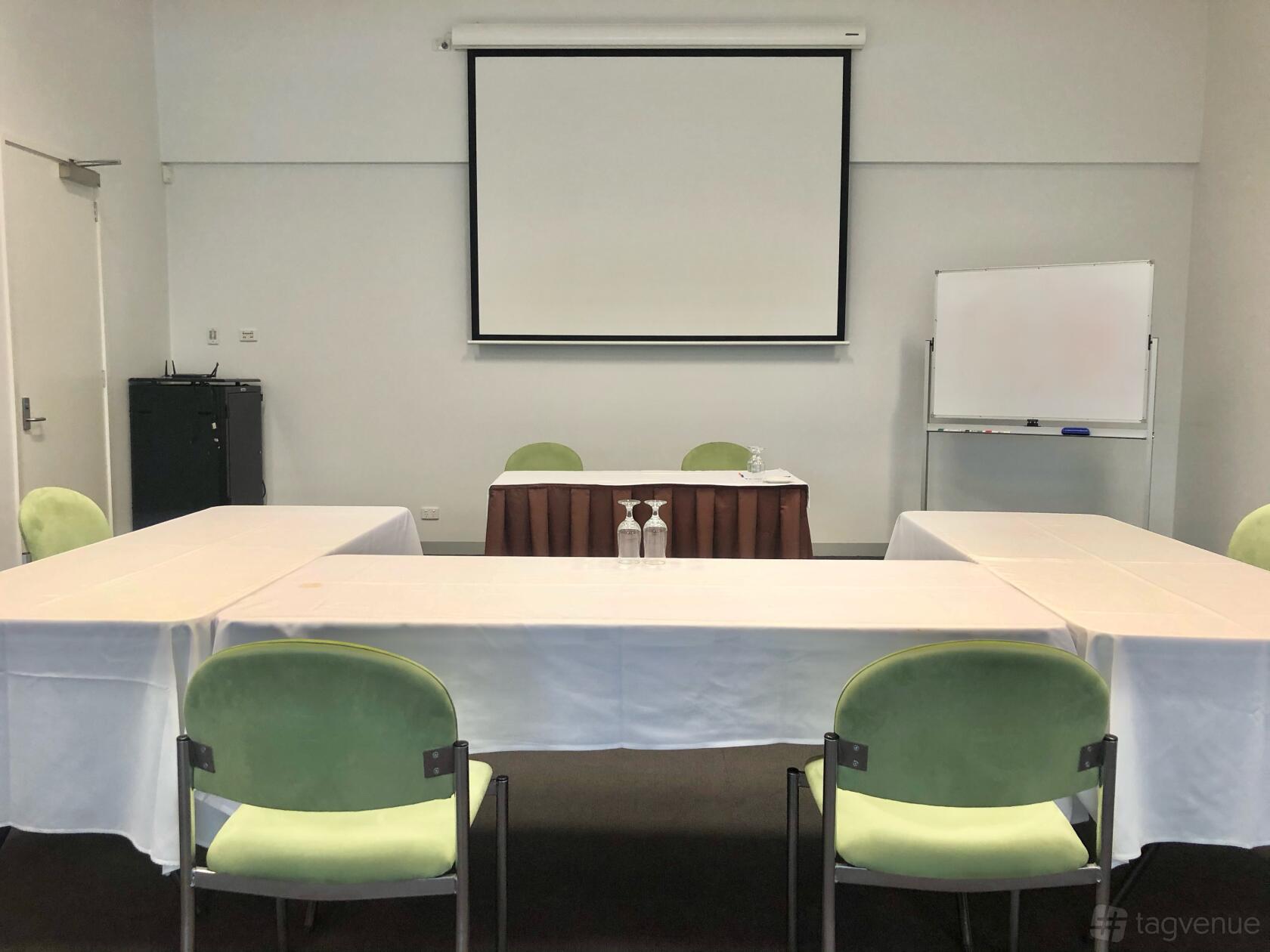 A meeting room with U-shaped tables covered in white linens, green chairs, a projector screen, and a whiteboard at Melbourne Polytechnic Conference Centre.