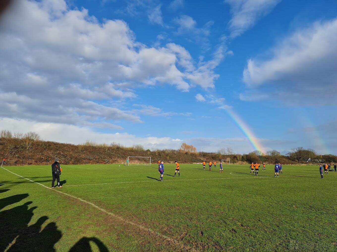 An open grass football pitch with players in action and a rainbow in the sky at Leicester Road Stadium.