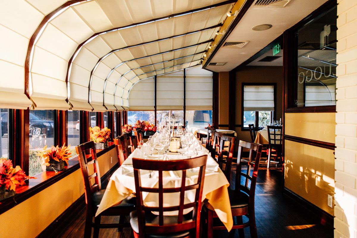 A restaurant with long tables set for dining, floor-to-ceiling windows with white blinds, and floral centerpieces at Il Luogo Ristorante.