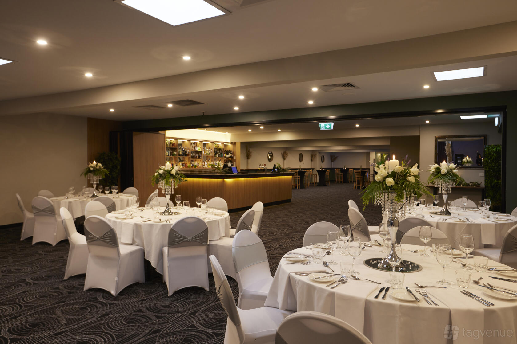 An event space in a pub with round tables dressed in white linens and floral centerpieces at Sandown Park Hotel.