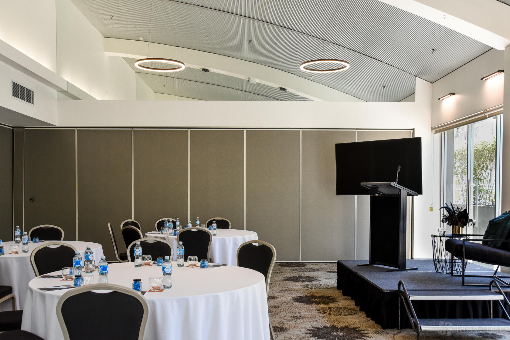 An event space with round tables, a stage with a podium, and natural light at The Terrace Hotel.