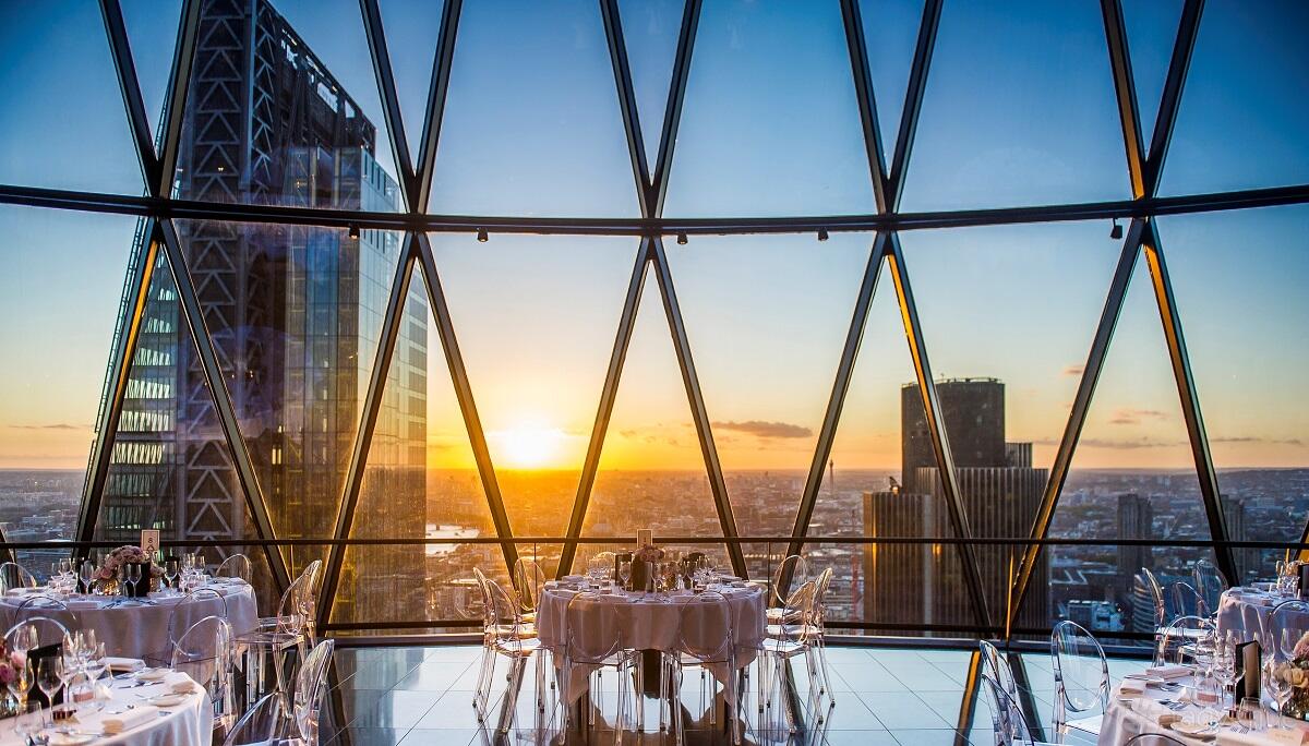 A restaurant with floor-to-ceiling glass windows, city skyline views, and round tables at Searcys at The Gherkin.