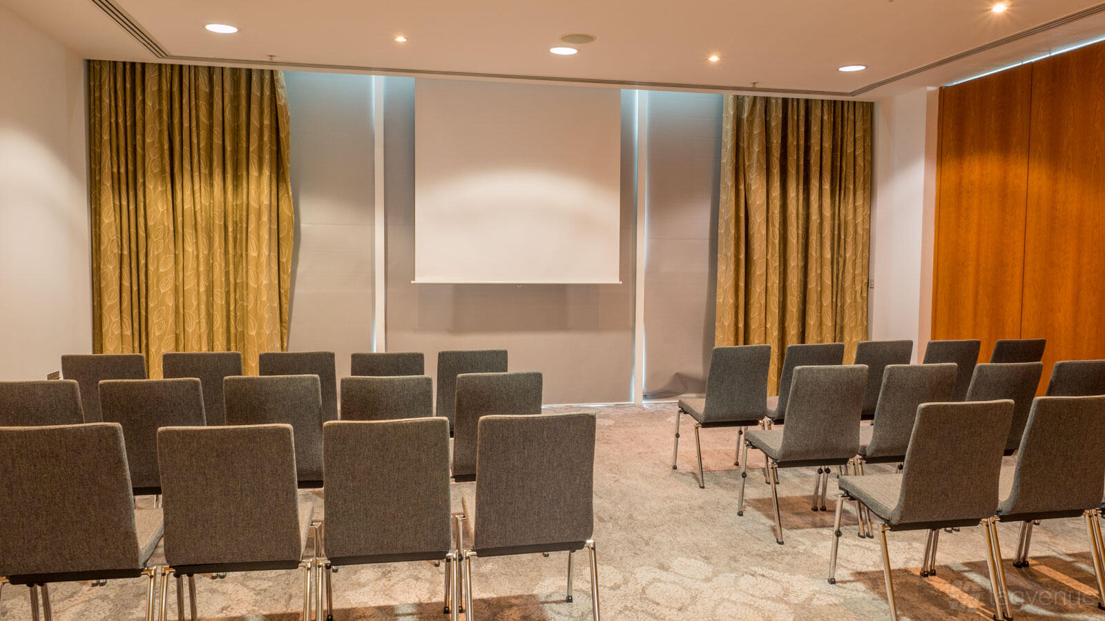 A meeting room with rows of gray chairs facing a projection screen and floor-to-ceiling curtains at The Lowry Hotel.