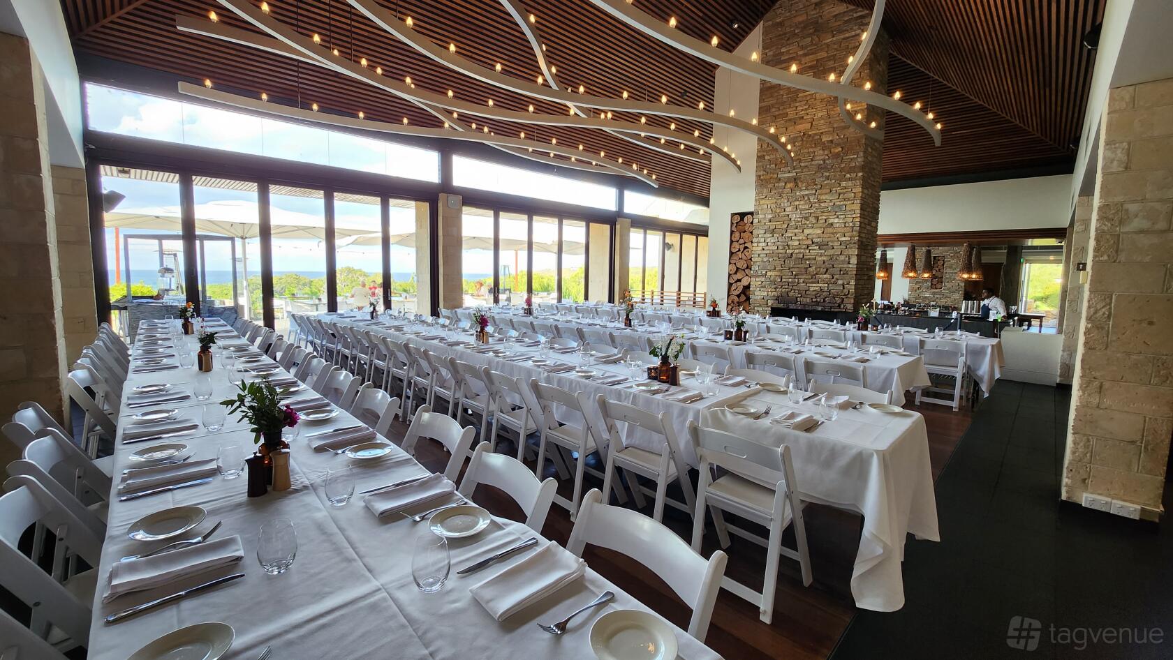 A hotel dining space with floor-to-ceiling windows, long banquet tables with white linens, and a stone fireplace at Pullman Bunker Bay.