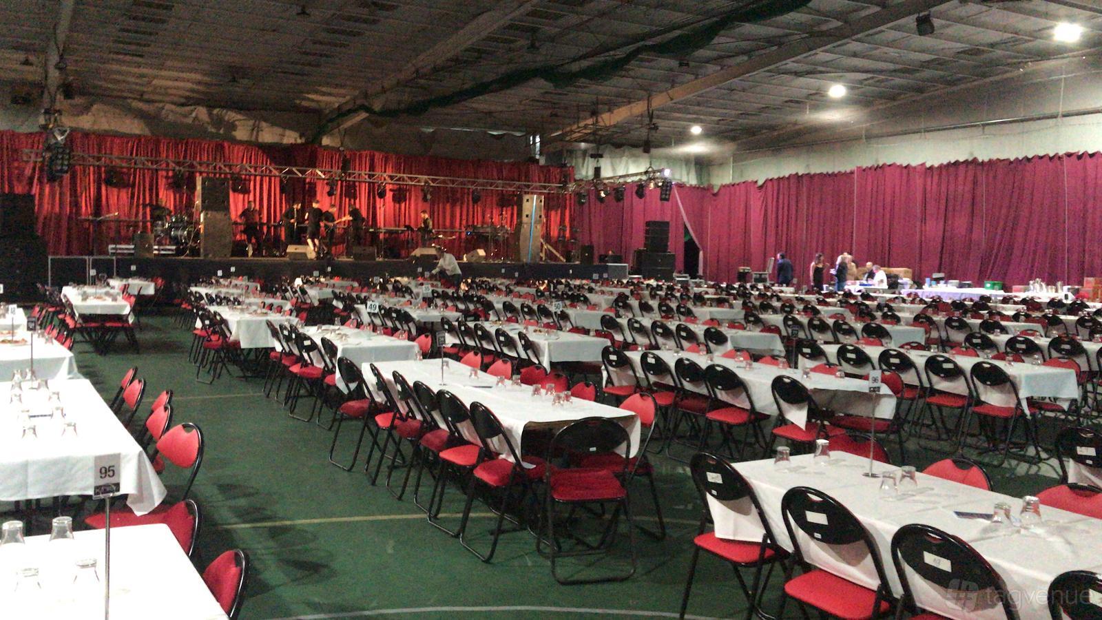 A sports hall with rows of banquet tables and chairs, stage setup, and red draped curtains at Westgate Indoor Sports.