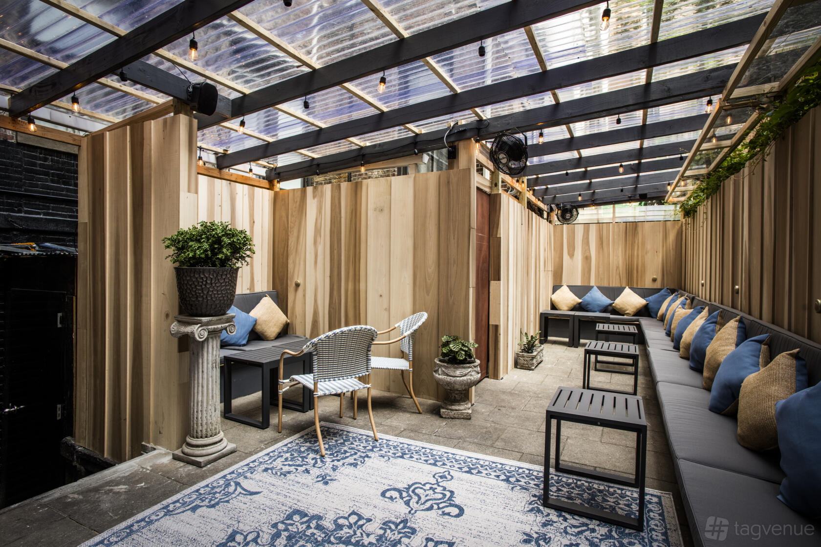 A covered outdoor lounge with wooden walls, string lights, long cushioned benches, and potted plants at Raines Law Room Chelsea.
