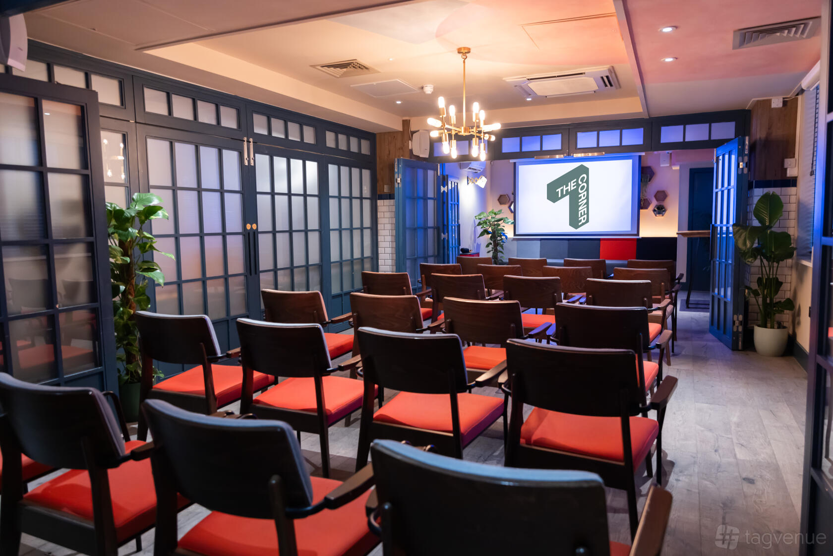 A hotel meeting room with rows of wooden chairs with red cushions, chandelier lighting, and a projector screen at The Corner London City.