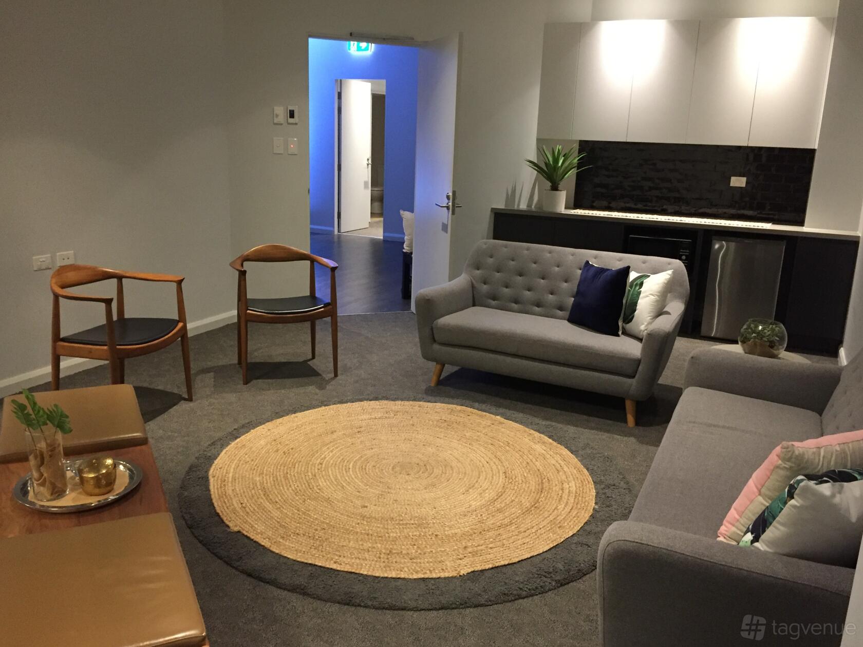 A meeting room with a gray sofa, wooden chairs, kitchenette, and a round woven rug at Camperdown Quarters.