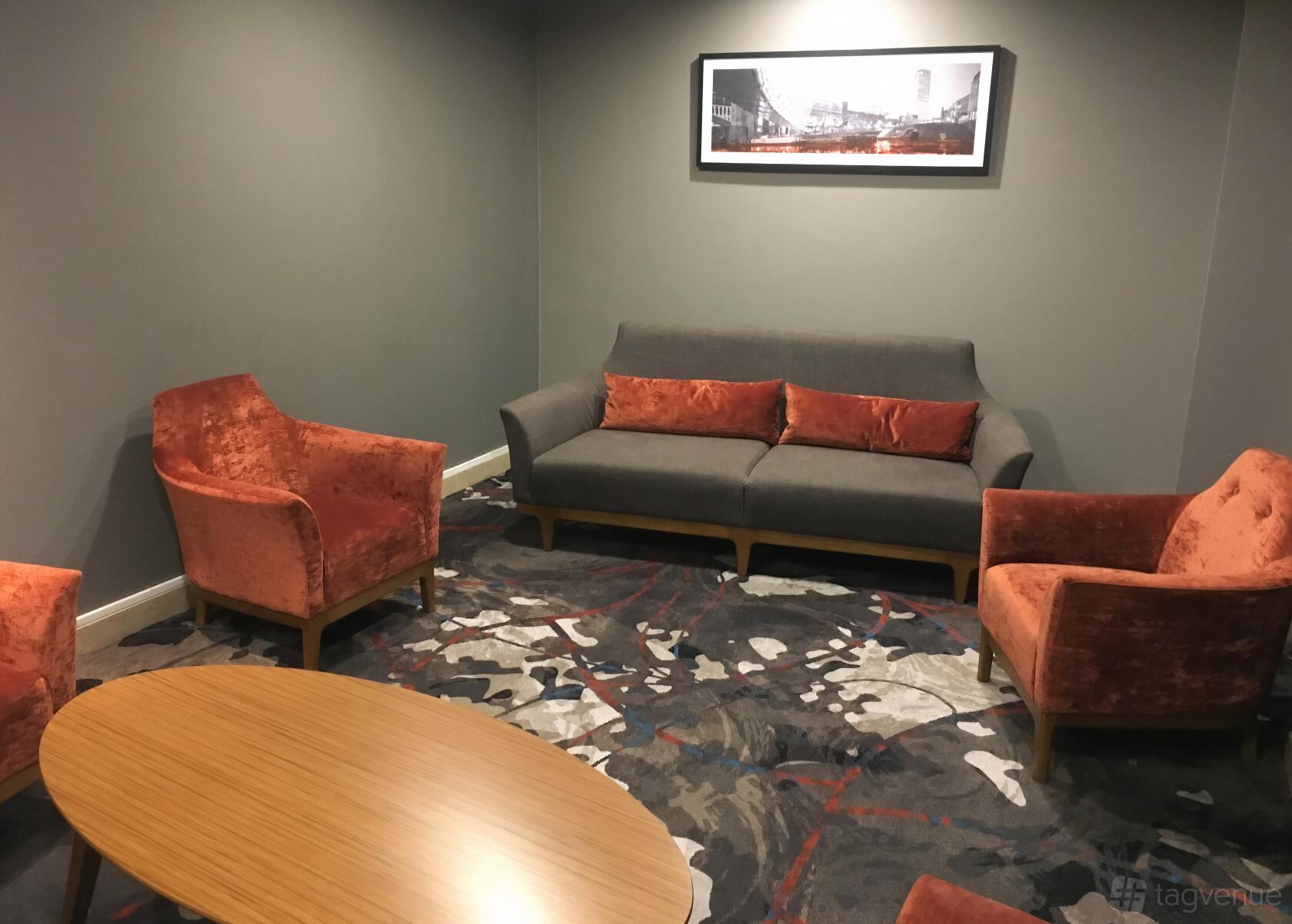 A meeting room with orange armchairs, a gray sofa, abstract patterned carpet, and a wooden coffee table at Clayton Hotel Manchester Airport.