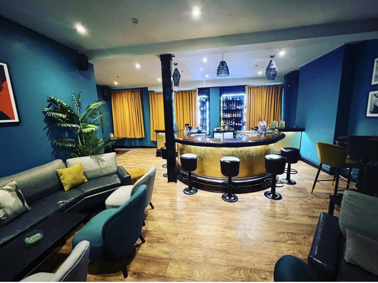 A pub lounge with a curved gold-lit bar, bar stools, teal walls, and mustard curtains at The Raven Tower Bridge.