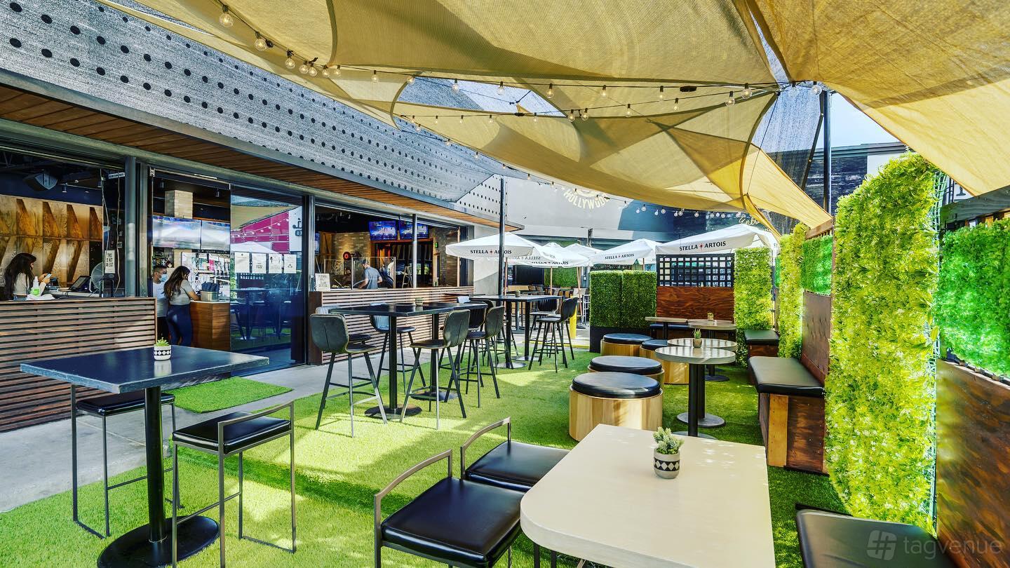 An outdoor restaurant patio with artificial grass, shaded sails, black tables, and hedge dividers at 3rd Base LA.