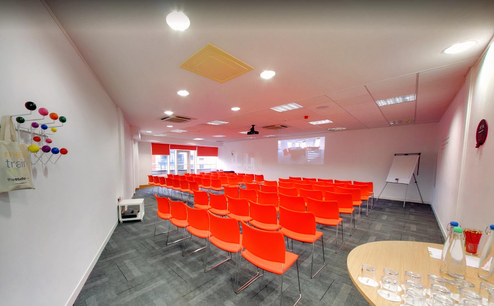 A meeting room with rows of orange chairs, projector, and flip chart at The Studio Leeds.