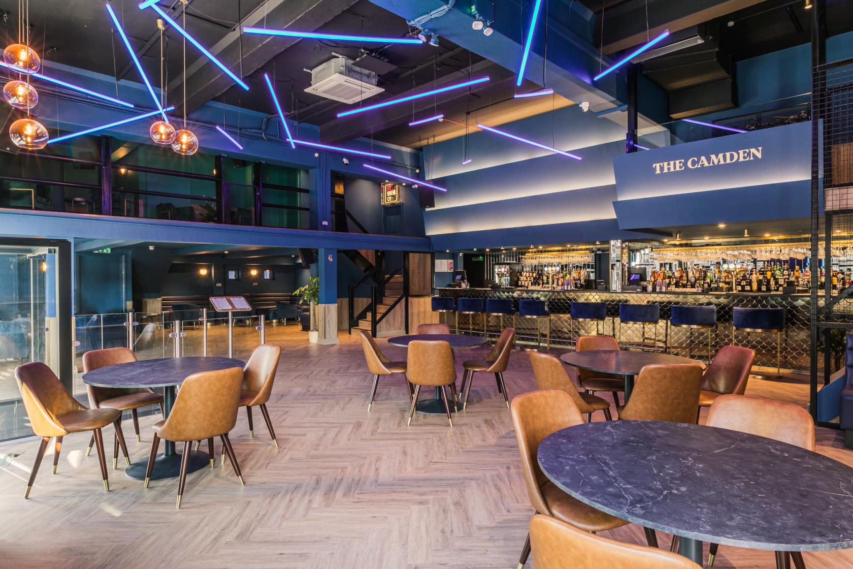 A bar with round marble tables, leather chairs, neon ceiling lights, and a mirrored bar counter at THE CAMDEN.