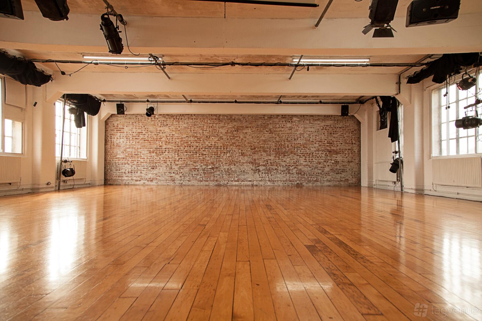 A rehearsal room with polished wooden floors, exposed brick wall, and overhead stage lighting at Chisenhale Dance Space.