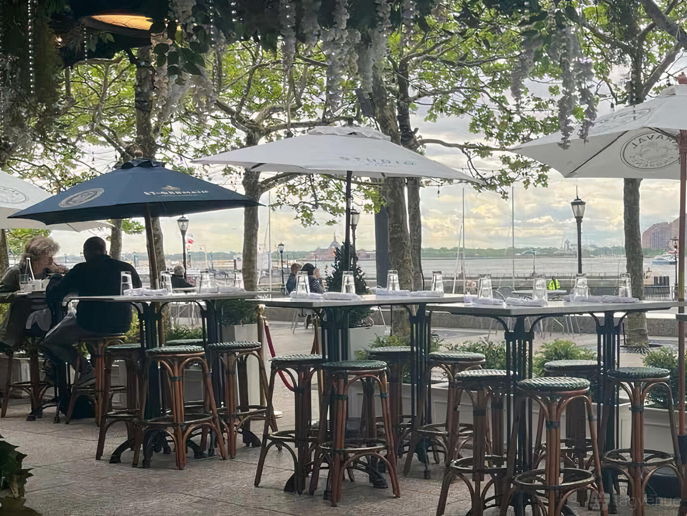 An outdoor bar area with high tables, stools, and umbrellas overlooking the waterfront at Le District.