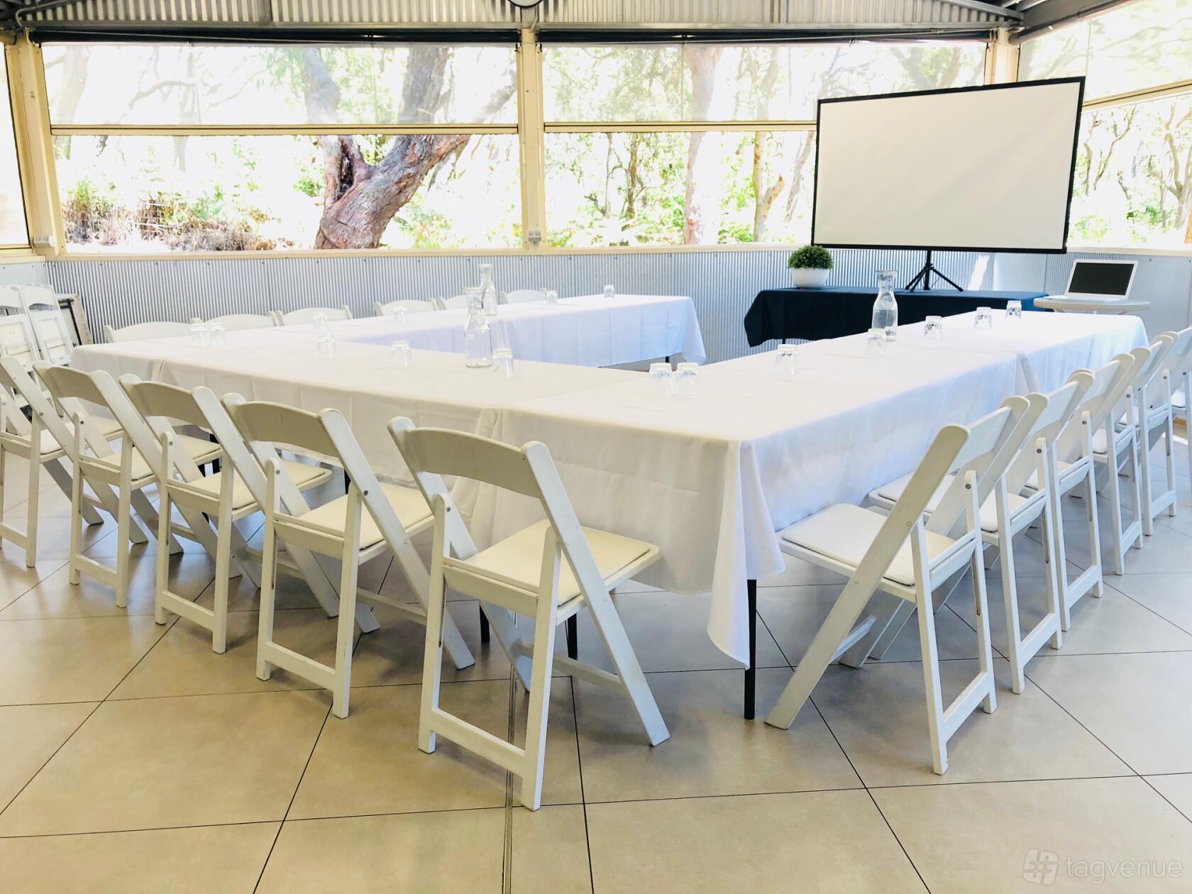 A meeting room with U-shaped tables covered in white linens, folding chairs, and a projector screen at The Retreat Port Stephens.