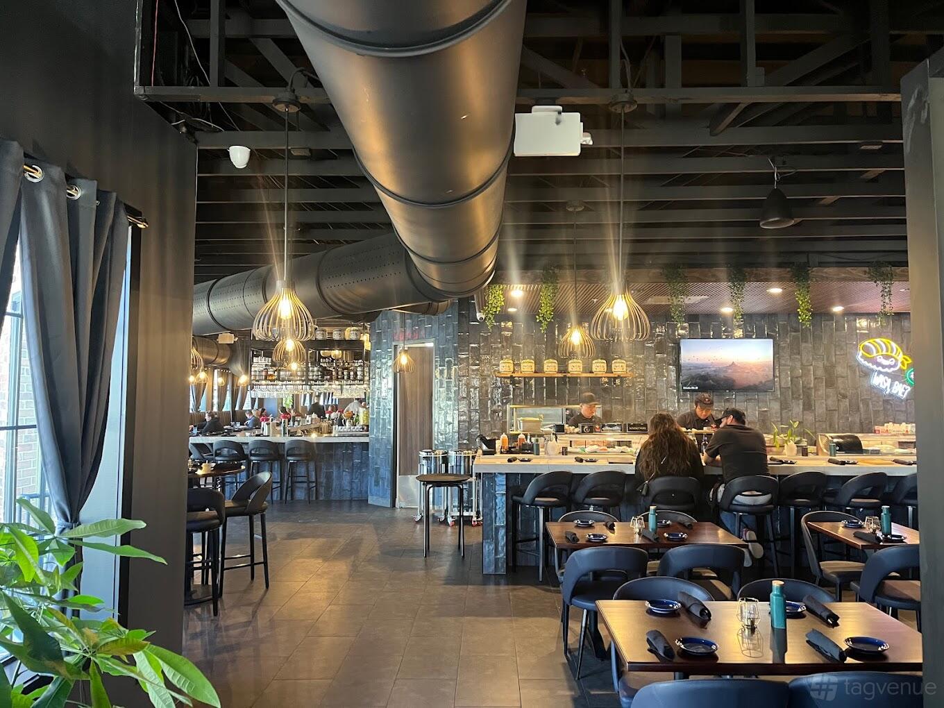 A restaurant with an open sushi counter, exposed ductwork ceiling, pendant lighting, and dark wood tables at Hush Sushi, Kitchen & Bar.