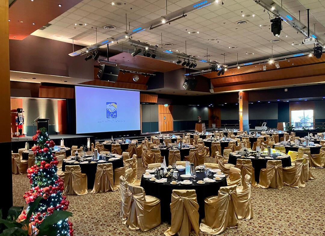 A ballroom with round tables draped in gold linens, banquet chairs, and a large projection screen at LCC Venues.
