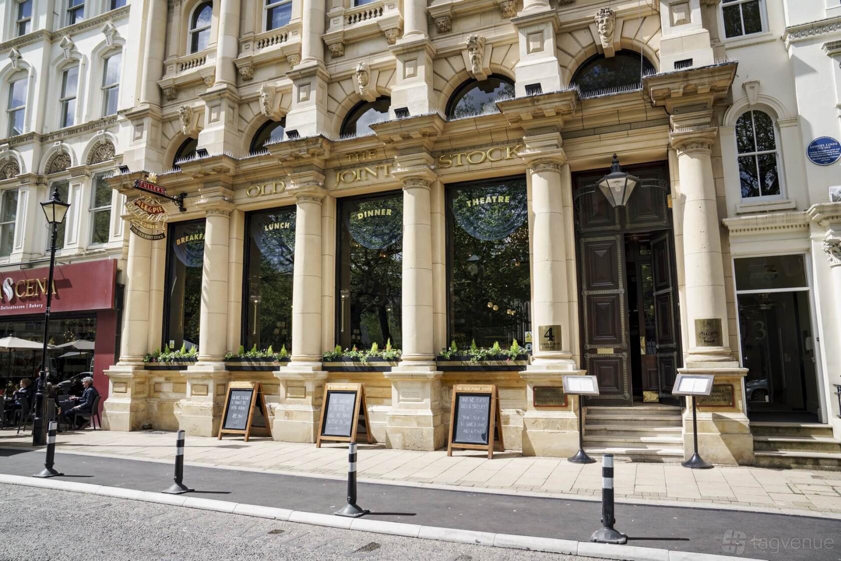 A pub with ornate stone facade, tall arched windows, column details, and menu boards outside at Old Joint Stock.