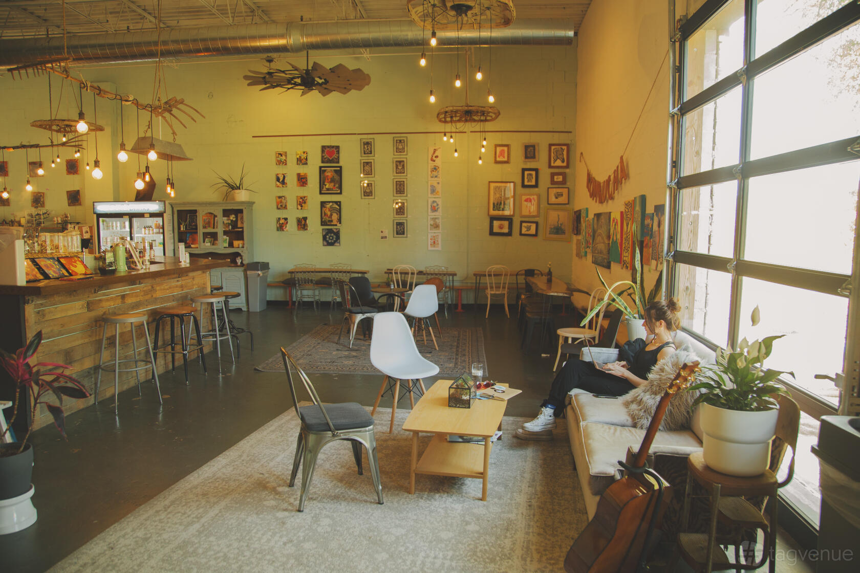 A taproom with eclectic wall art, hanging pendant lights, indoor plants, and mixed seating at The Cultured South Taproom.