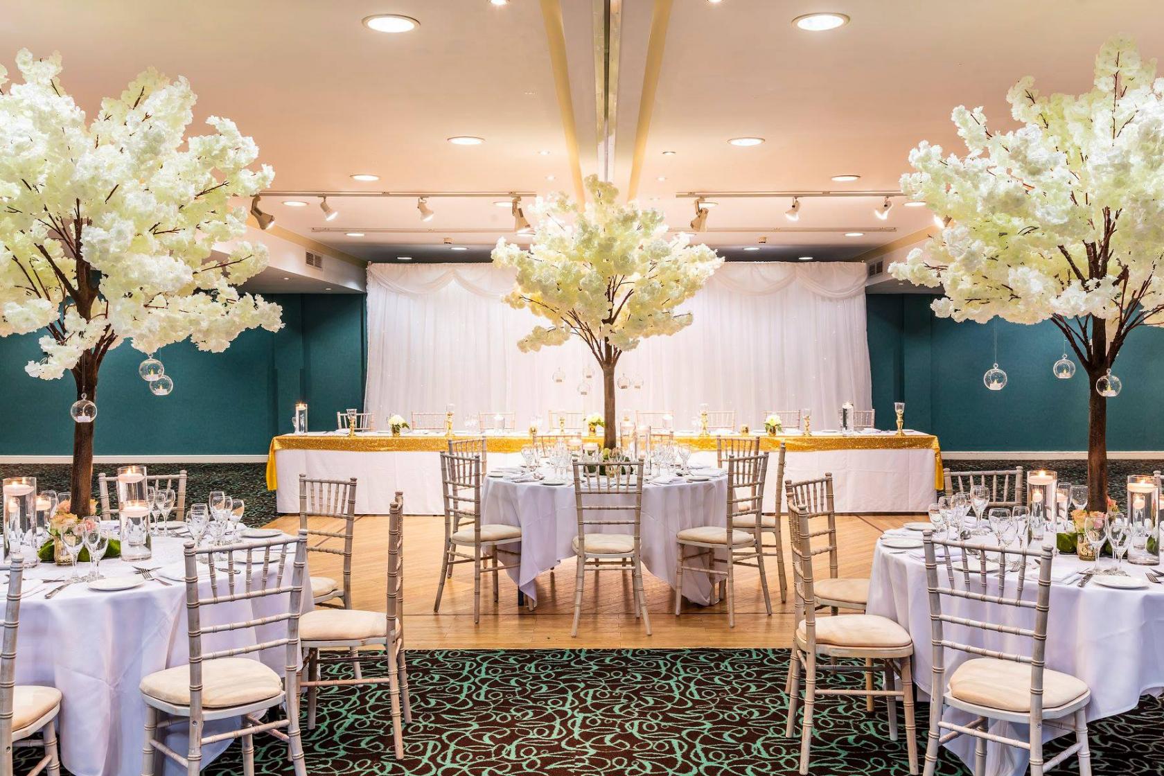 A hotel ballroom with round tables, white chairs, and large artificial blossom trees at Castle Green Hotel.