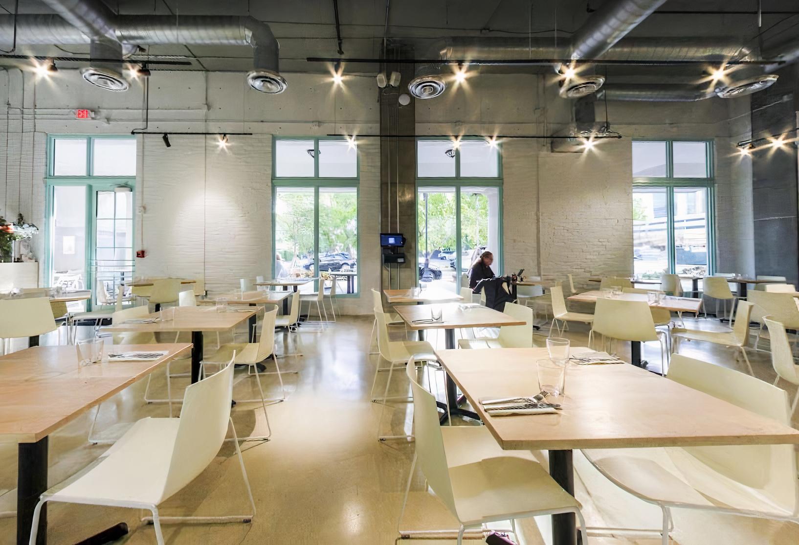 A restaurant with large windows, light wood tables, white chairs, and exposed ductwork at Bachour.