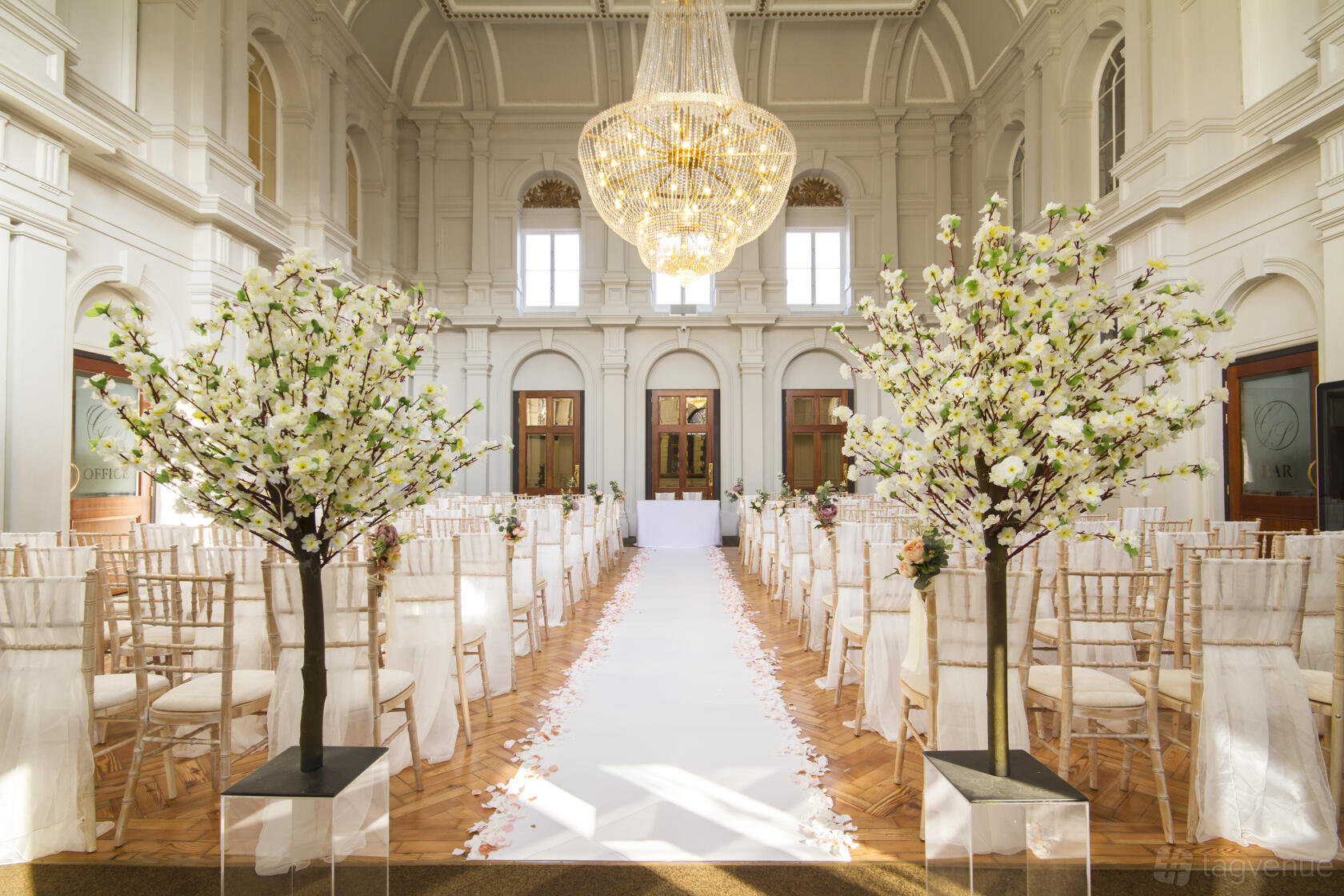 A historic event hall with high vaulted ceilings, a grand chandelier, and rows of chairs along a petal-lined aisle at The Grand Station.