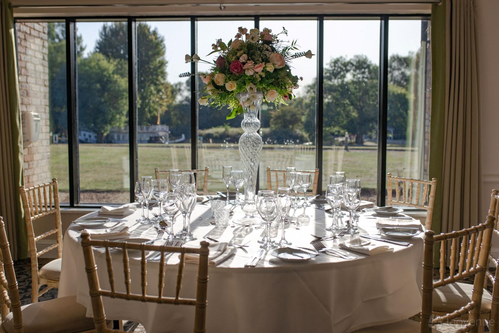 An event space with a round table set with glassware and a floral centerpiece by large windows at Voco Oxford Thames.