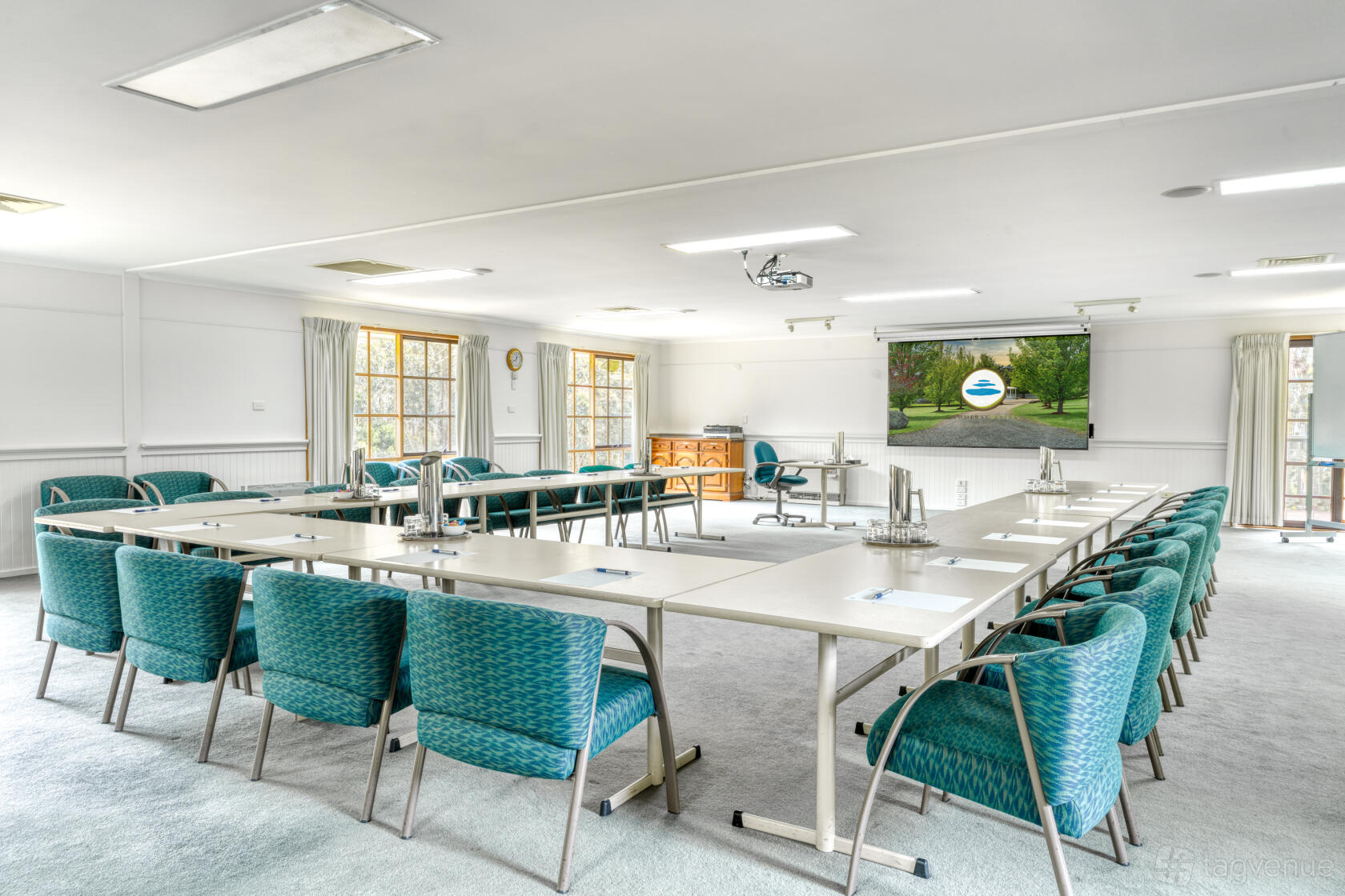 A meeting room with U-shaped tables, teal cushioned chairs, natural light, and a projector screen at Cammeray Waters Function Centre.