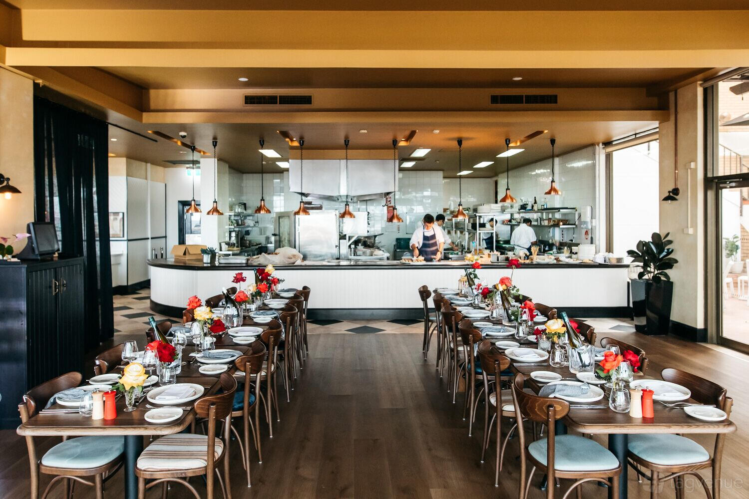 A dining room with two long tables set with floral centerpieces, wooden chairs, and an open kitchen at Bistro Manly.