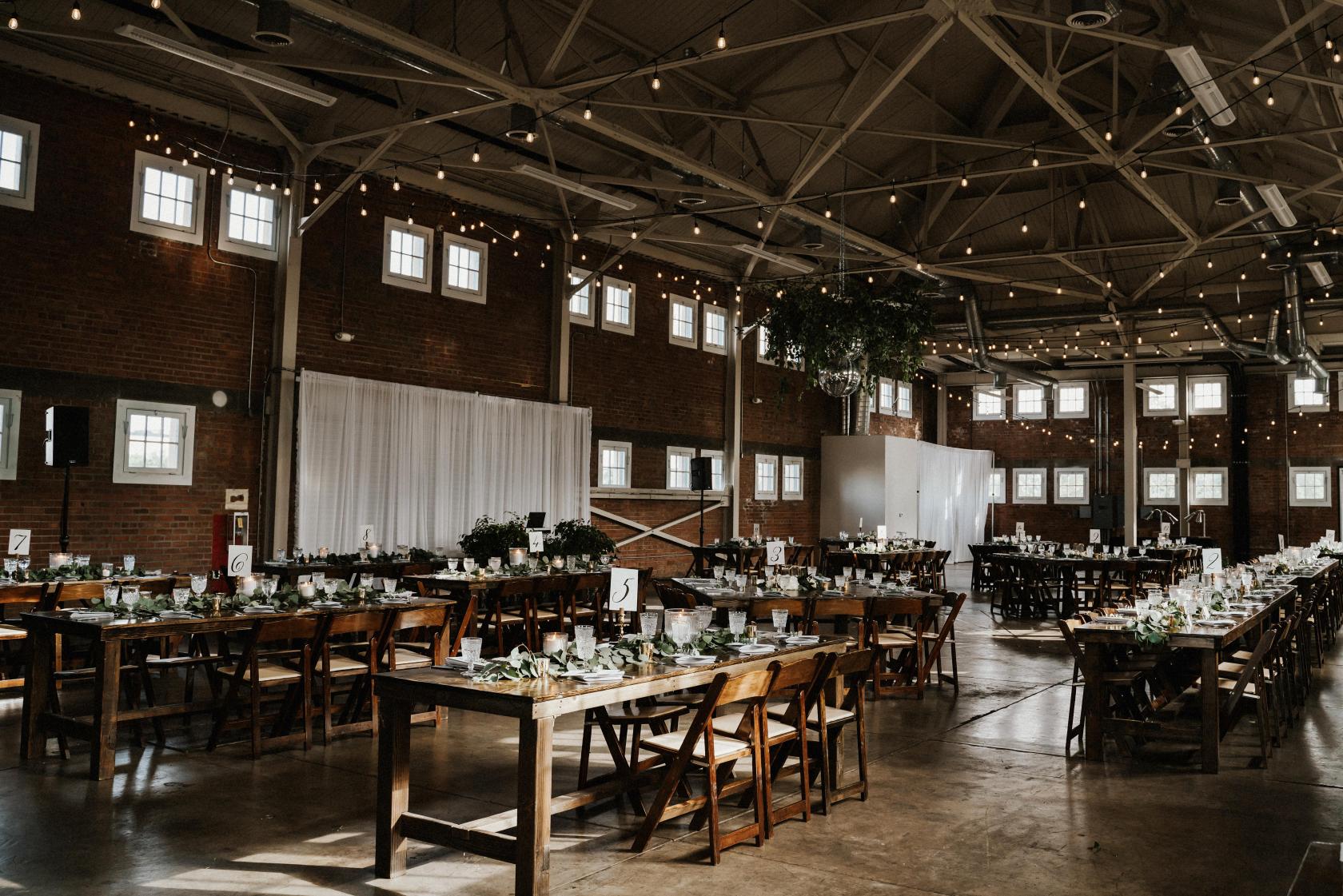 A hall with exposed brick walls, high industrial ceilings, string lights, and banquet tables at BRICK.
