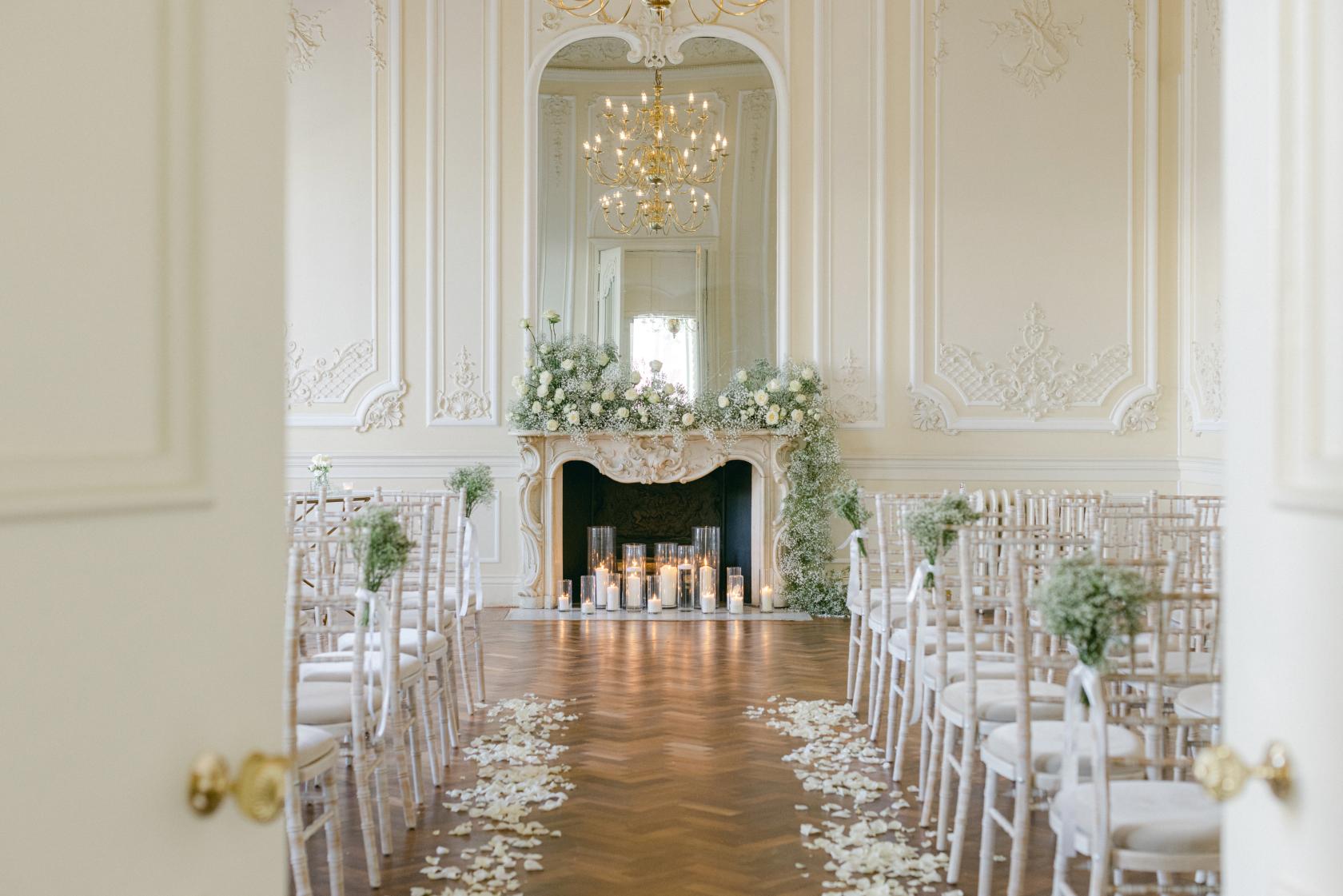 An event venue with ornate white walls, chandelier lighting, and a floral-decorated fireplace at {10-11} Carlton House Terrace.