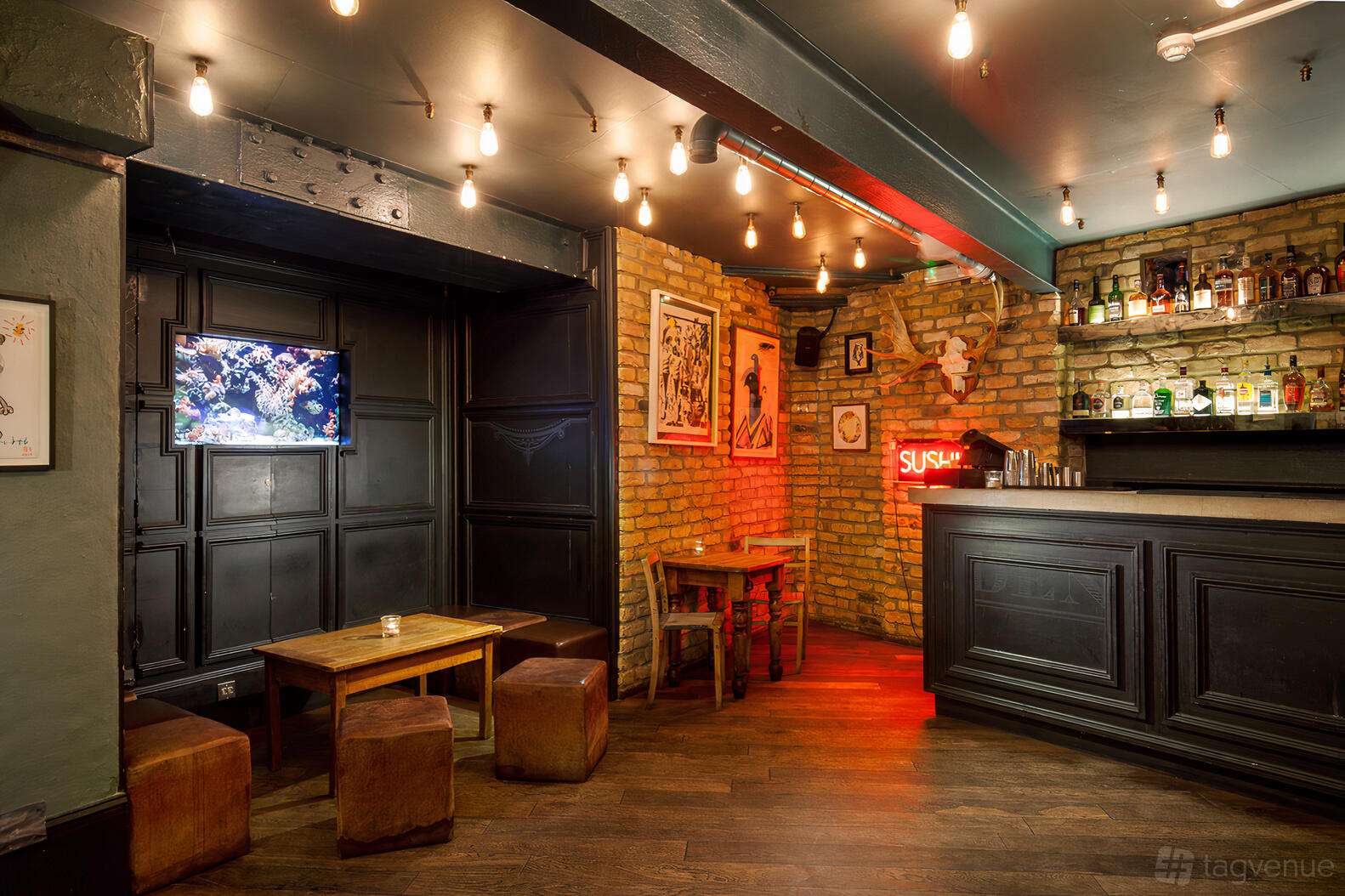 A bar basement with exposed brick walls, warm lighting, a mounted TV, and a black wood bar at The Alice House West Hampstead.