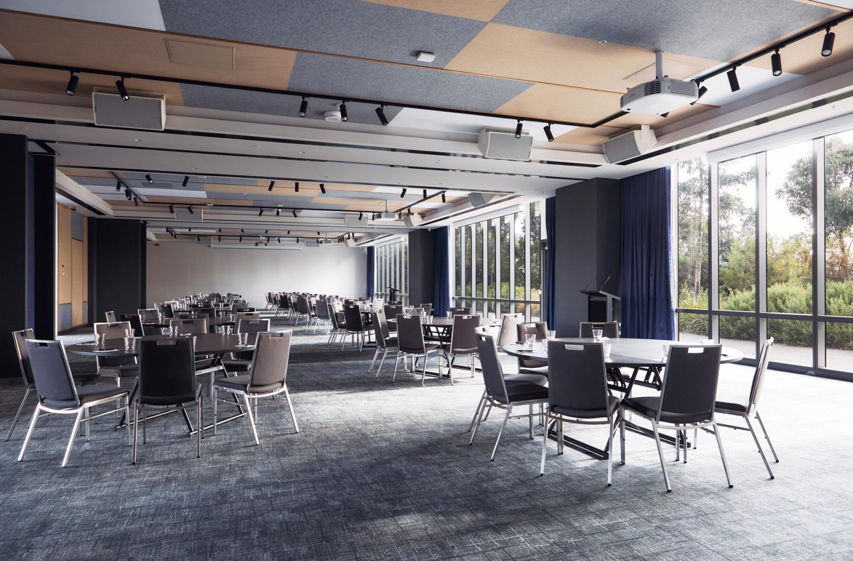 A hotel ballroom with round tables, black chairs, large floor-to-ceiling windows, and natural lighting at Mantra Melbourne Epping.