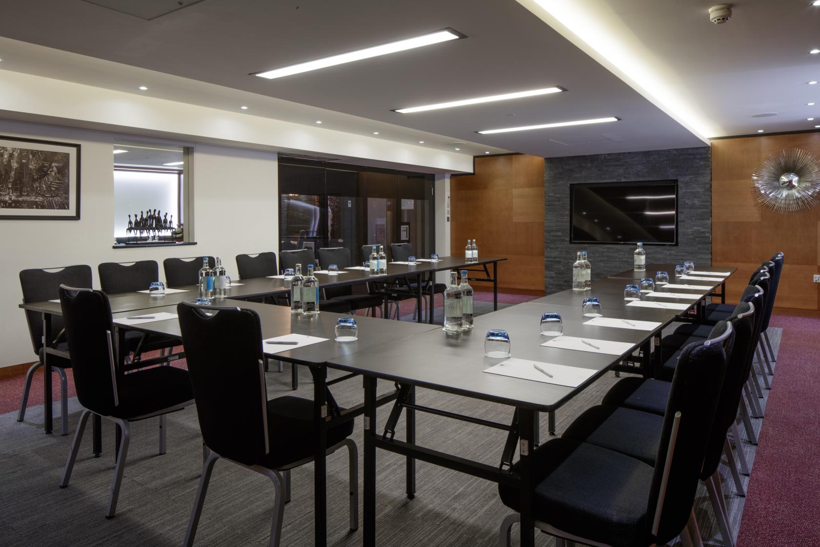 An event space with U-shaped tables, black chairs, bottled water, and a large wall-mounted screen at DoubleTree by Hilton London Greenwich.