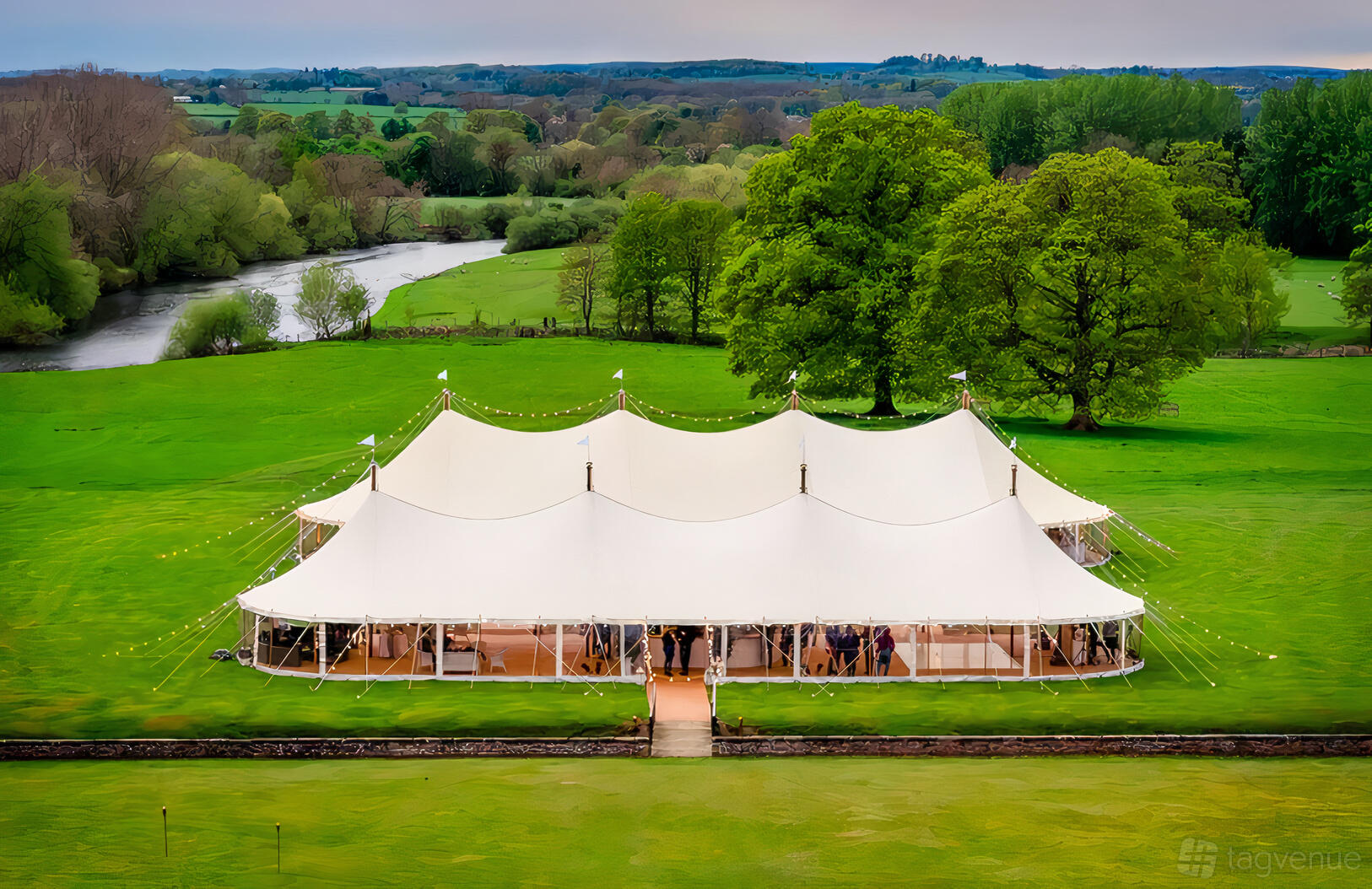 An outdoor venue with a large white marquee tent set on a green lawn by a river at Peacock and Bow.
