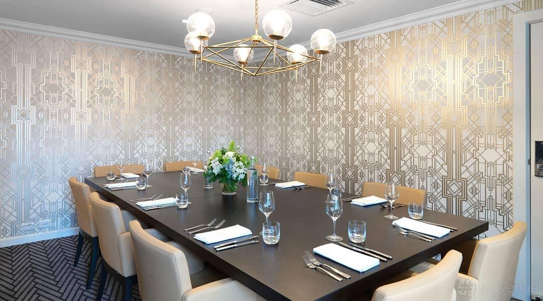 A dining room with geometric wallpaper, a rectangular table set with cutlery, and a modern chandelier at Hotel Kurrajong Canberra.