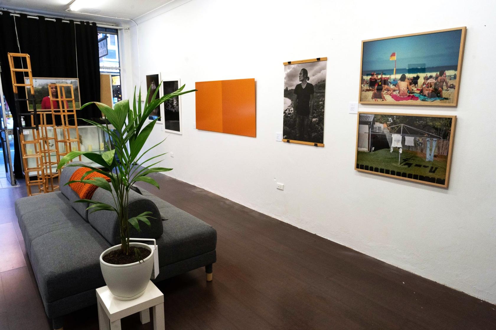 A gallery with framed art prints on white walls, dark wood floors, and a gray sofa with a potted plant at LUNA STUDIO SYDNEY.