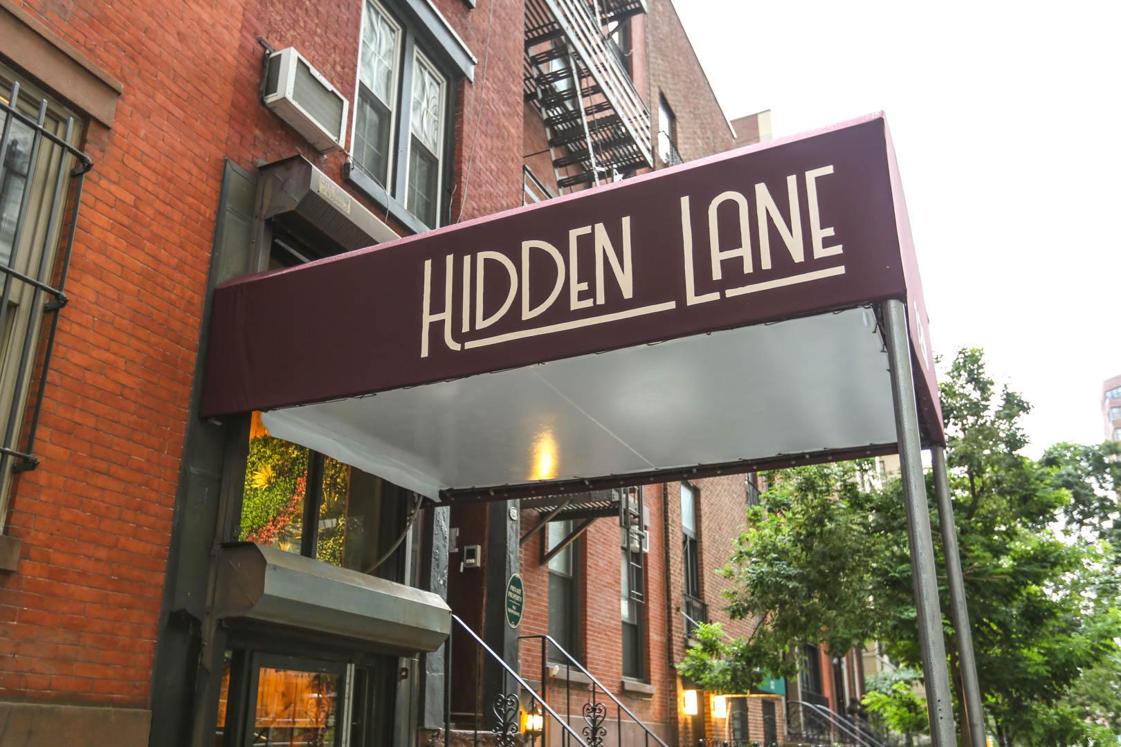A pub entrance with a maroon awning labeled 'Hidden Lane' and adjacent brick building at Hidden Lane Bar.