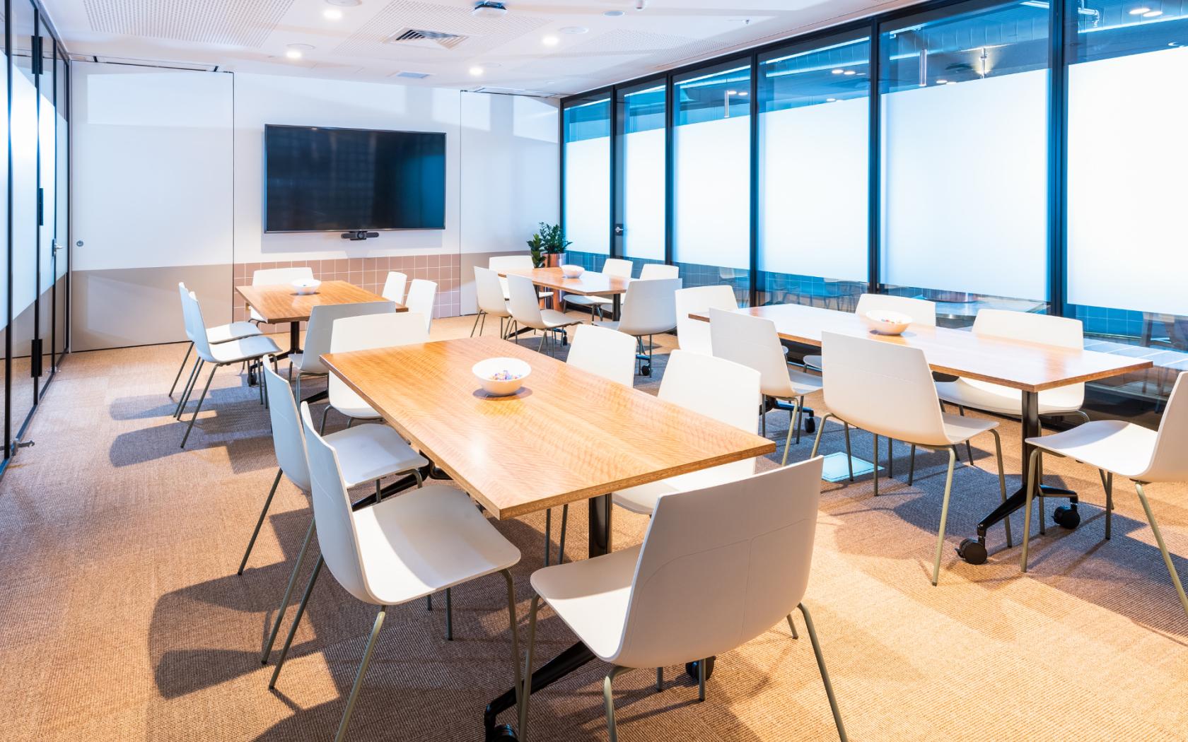 A meeting room with large wooden tables, white chairs, a wall-mounted screen, and floor-to-ceiling windows at Flex by ISPT at Central Plaza.