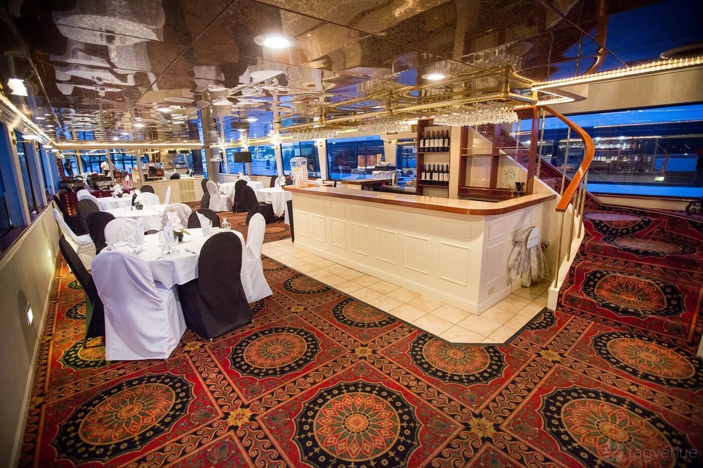 An event venue with floor-to-ceiling windows, patterned carpet, a central bar, and round dining tables at Chardonnay.